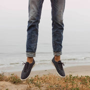 Men's Hermosa Sneaker in Slate Navy canvas, worn with cuffed jeans, jumping on a beach