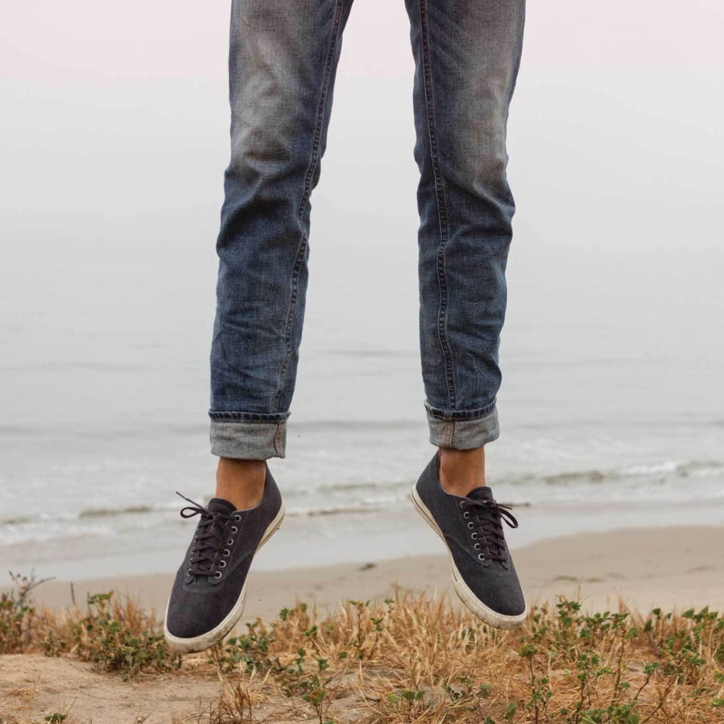 Men's Hermosa Sneaker in Slate Navy canvas, worn with cuffed jeans, jumping on a beach