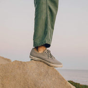 Men's Hermosa Sneaker in Tin Grey canvas, worn with green pants, foot resting on a coastal rock