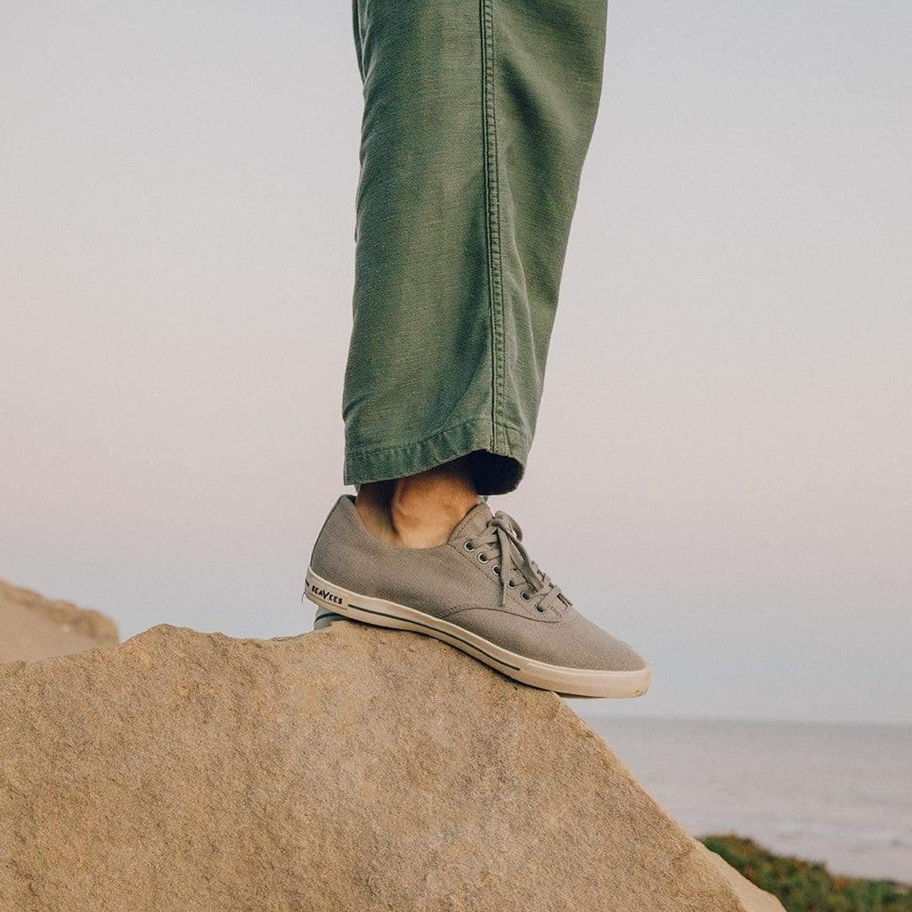 Men's Hermosa Sneaker in Tin Grey canvas, worn with green pants, foot resting on a coastal rock