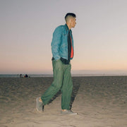 Men's Hermosa Sneaker in Tin Grey canvas, worn with green pants by a person standing on a beach at dusk