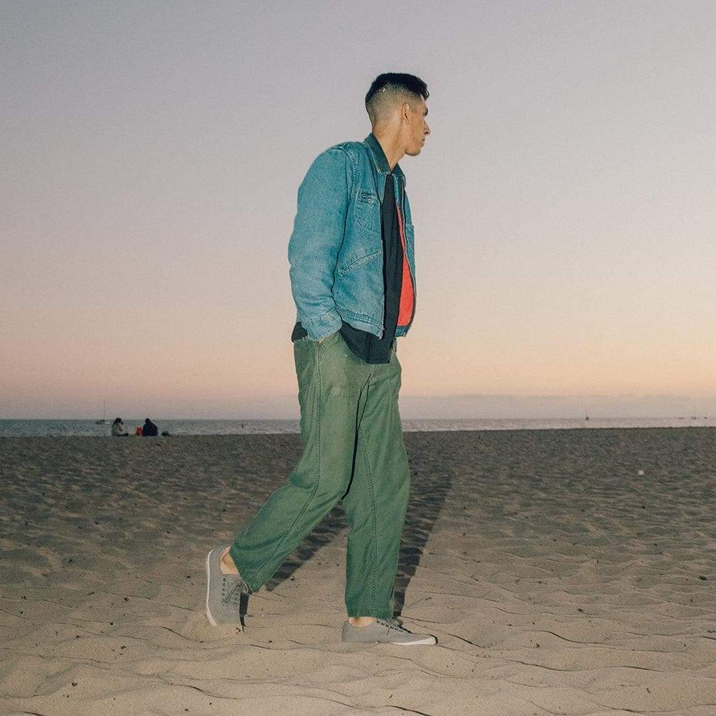 Men's Hermosa Sneaker in Tin Grey canvas, worn with green pants by a person standing on a beach at dusk