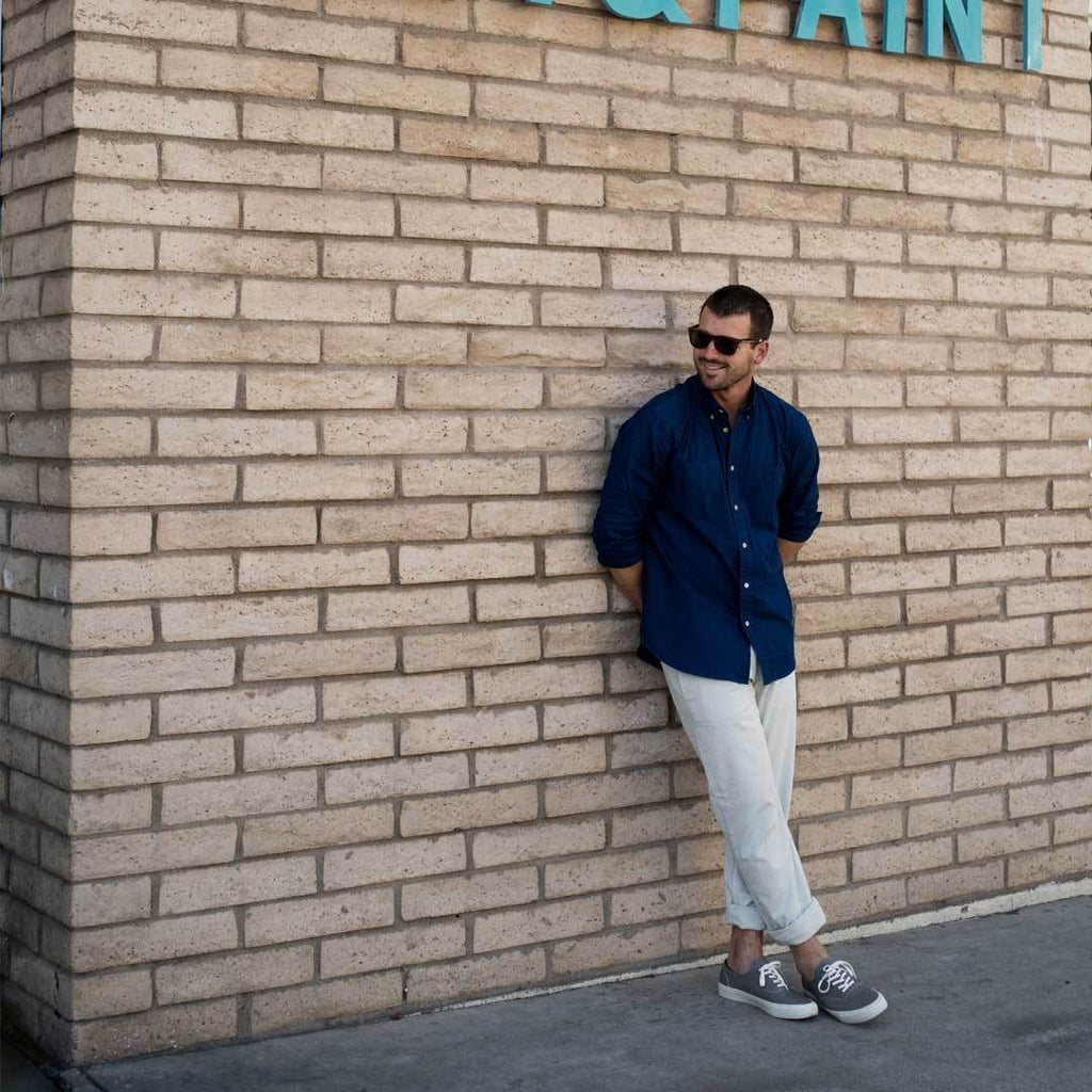 Men's Legend Sneaker in Granite Grey canvas, worn by a person leaning against a brick wall in a navy shirt