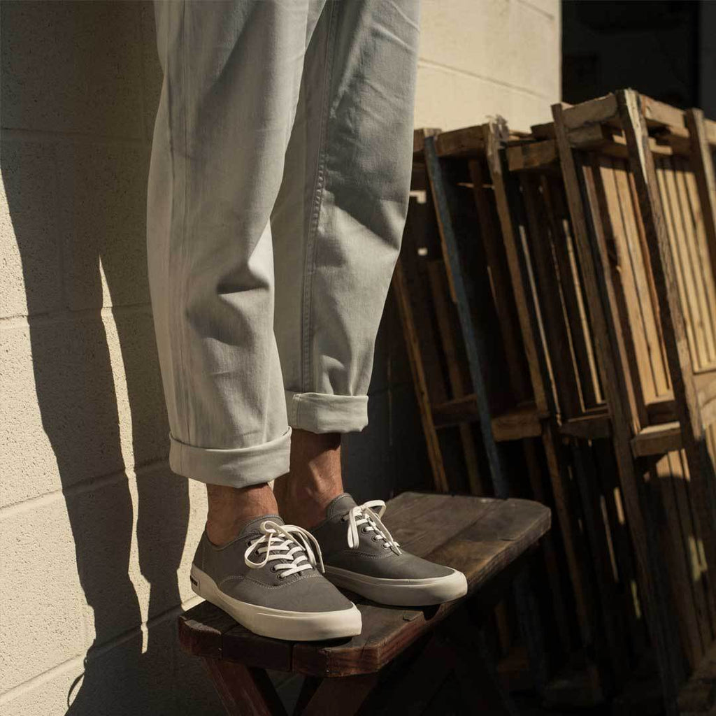 Men's Legend Sneaker in Granite Grey canvas, worn with gray pants, seated beside wooden crates