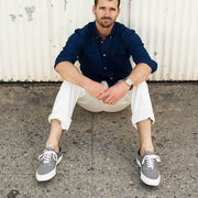 Men's Legend Sneaker in Granite Grey canvas, worn with white pants, seated on pavement in a navy shirt