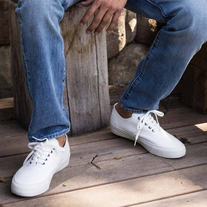 Men's Legend Sneaker in Bleach canvas, worn with jeans, seated on a weathered wooden post