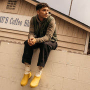 Men's Bodega Clog in Calendula rubber, worn with cream socks by a person seated on a concrete wall