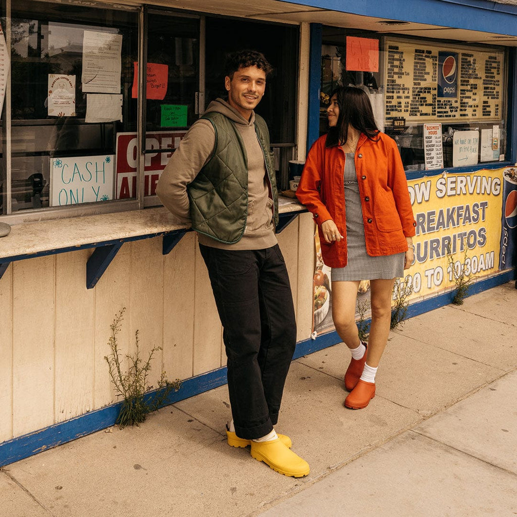 Men's Bodega Clog in Calendula rubber, worn with an olive jacket by a person standing outside a food stand