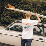 Person in a SeaVees white t-shirt carrying a surfboard beside a car