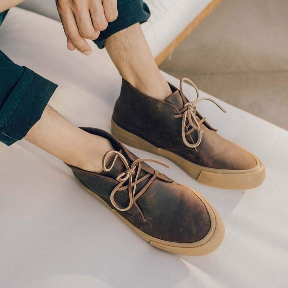 Men's Maslon Desert Boot in Kona leather, worn by a person seated with feet crossed on a white surface