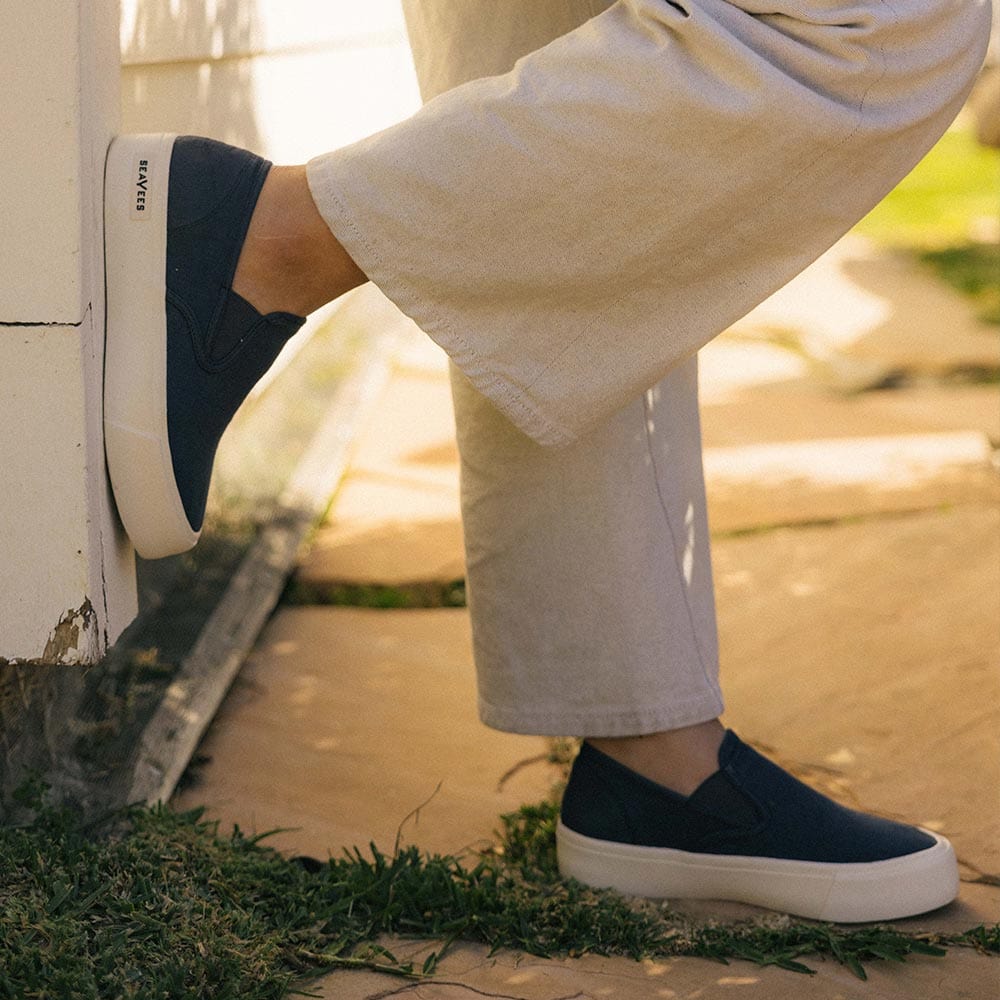 Women's Baja Slip On in True Navy canvas, worn with cream pants, one foot raised outdoors
