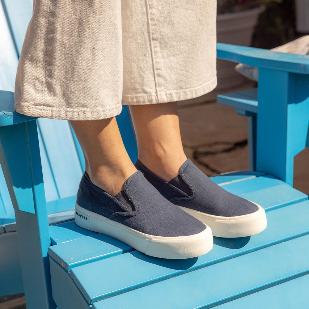 Women's Baja Slip On in True Navy canvas, worn with cream pants, seated on a blue Adirondack chair