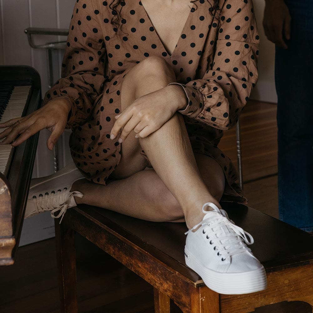Women's Monterey Sneaker in Bleach canvas, worn with a polka dot dress, seated on a wooden bench