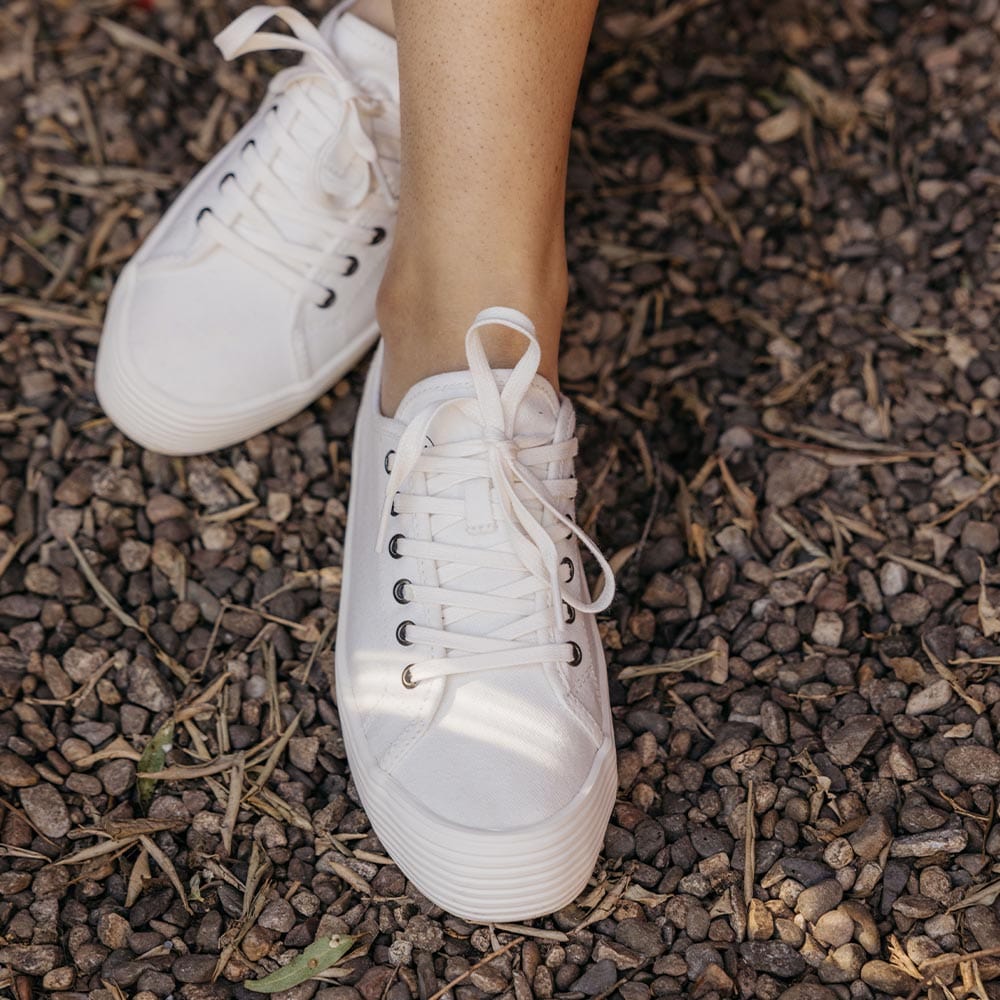 Women's Monterey Sneaker in Bleach canvas, worn with bare ankles on dark mulch, close up
