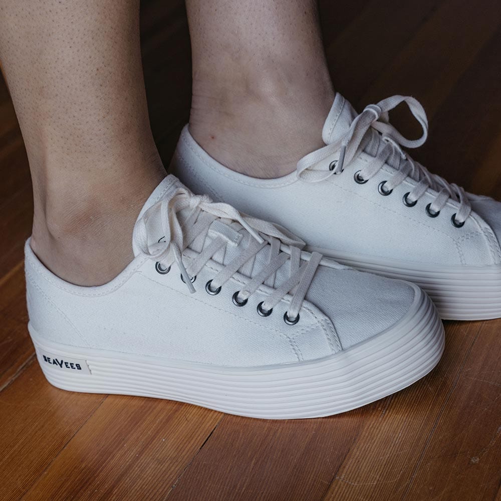 Women's Monterey Sneaker in Bleach canvas, worn with bare ankles on a wood floor, side view