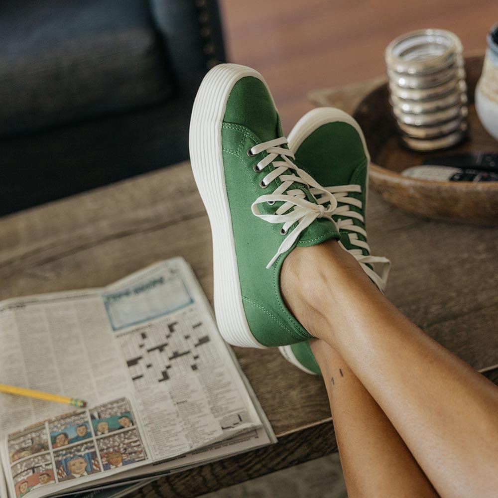 Women's Monterey Sneaker in Cactus canvas, worn with crossed legs beside a crossword puzzle