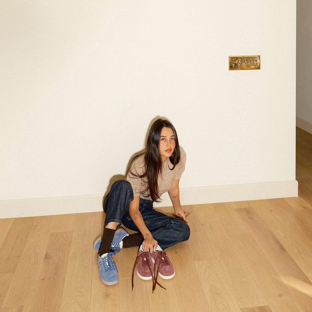 Women's Clipper Sneaker in Cornflower Blue suede, worn by a person seated on a wood floor holding a burgundy pair