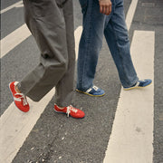 Women's Parsons Low Profile Trainer sneaker in Ribbon Red nylon and suede, worn with gray pants, walking across a crosswalk