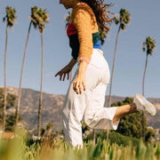 Women's Monterey Sneaker in Bleach canvas, worn by a person mid-jump in white pants among palm trees