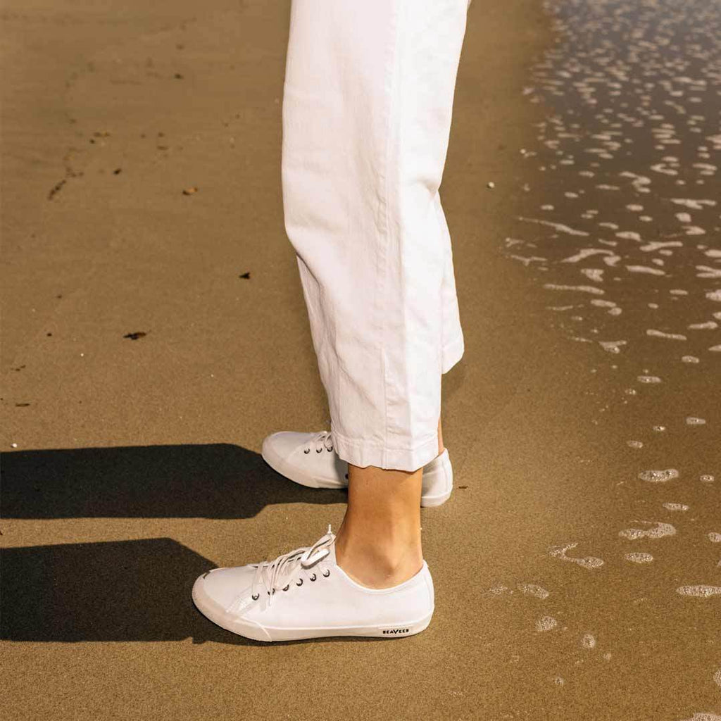 Women's Monterey Sneaker in Bleach canvas, worn with white pants on a sandy beach