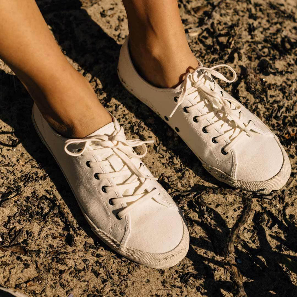 Women's Monterey Sneaker in Bleach canvas, worn bare-ankled on rough sandy ground, close up