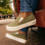 Women's Baja Slip On in Surplus Green canvas, close-up worn with cuffed jeans on a brick sidewalk