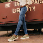 Women's Baja Slip On in Surplus Green canvas, worn with jeans in front of a red train car