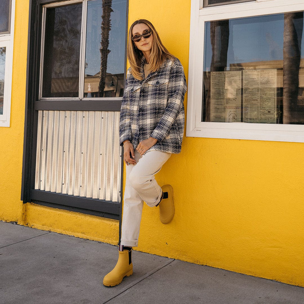Women's Bolinas Off Shore Boot in Calendula rubber, worn with white pants by a person standing against a yellow wall