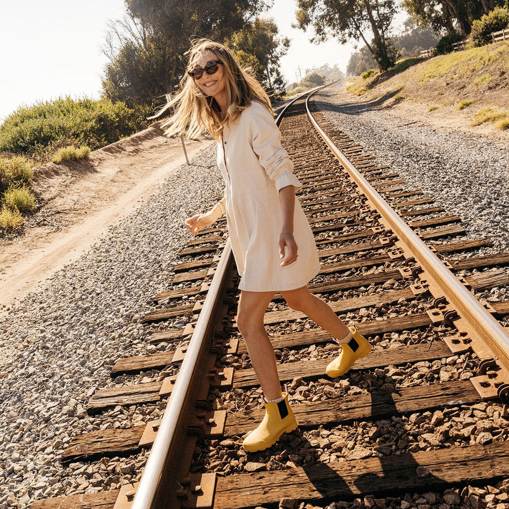 Women's Bolinas Off Shore Boot in Calendula rubber, worn with a white dress by a person walking along railroad tracks